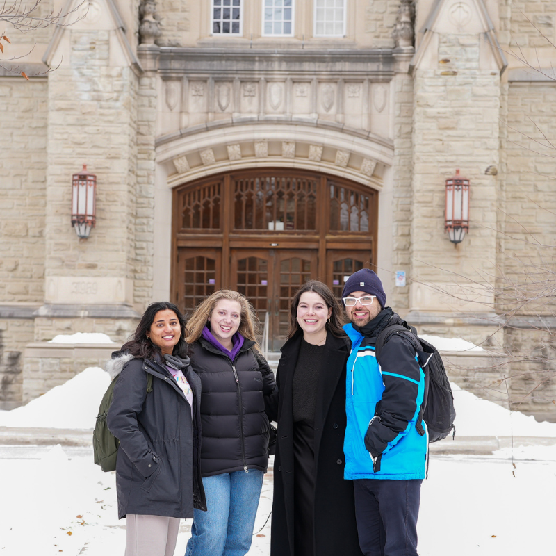 Four students in the snow stand outside an academic building.